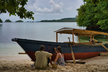 Back view of unrecognizable couple sitting on sandy beach at seaside and writing on boat with red paint