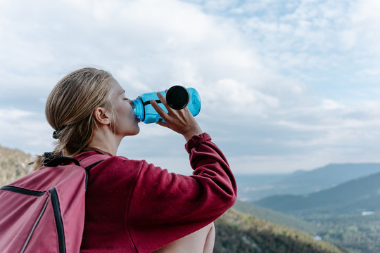 Side view of young female backpacker sitting on stone against green forested hill and drinking water while resting during hiking in Navacerrada in Spain
