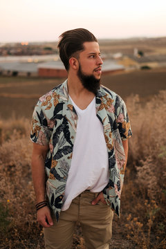 Thoughtful bearded male in casual wear standing near growing grass illuminated by sun at sundown in summer in countryside and looking away