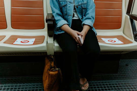 Crop unrecognizable female in casual outfit with smartphone in hands sitting on passenger seat in underground transport with markings for keeping distance during coronavirus pandemic