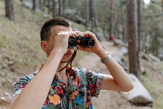 Male tourist in colorful shirt standing on slope of mountain covered with coniferous forest and observing environment with binoculars during hiking in Navacerrada in Spain