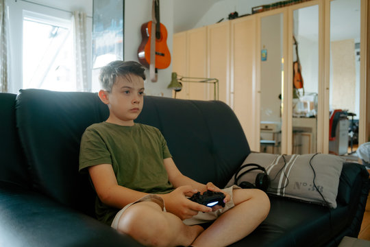 Concentrated little boy in casual clothes sitting on sofa with crossed legs and playing video game with joystick