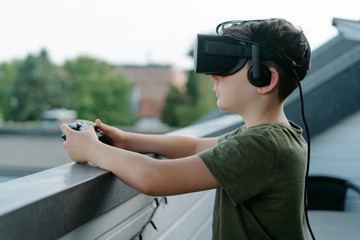 Curious little boy in t shirt standing on balcony alone and looking away using VR goggles and joystick
