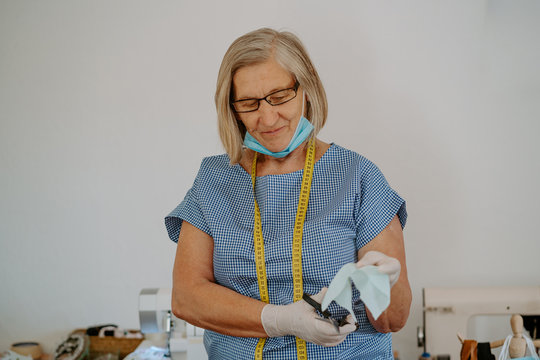 Craftswoman in latex gloves standing cutting piece of cloth with scissors for protective mask for coronavirus prevention at workplace
