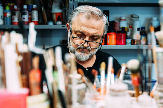 focused mature artist in eyeglasses working behind collection of paintbrushes of different sizes and bottles with paint in creative studio