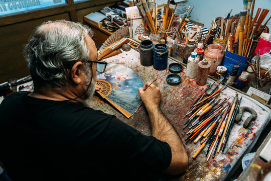 Back view from above faceless male painter using paintbrush to draw on hand fan while sitting at messy table and working in creative workshop