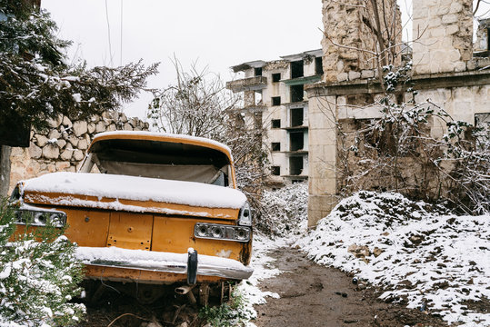 Abandoned weathered automobile covered with snow parked in countryside near ruined stone buildings in winter