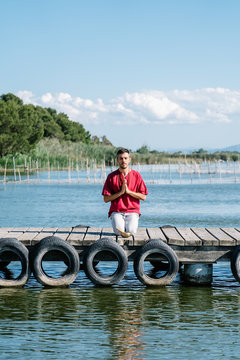 Peaceful man in comfy clothes sitting on wooden pier of sea and meditating in Hero yoga pose with namaste gesture