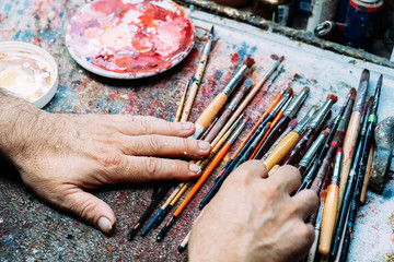 Crop concentrated mature male choosing paintbrush for mixing blue and white paints in palette while working in creative workshop