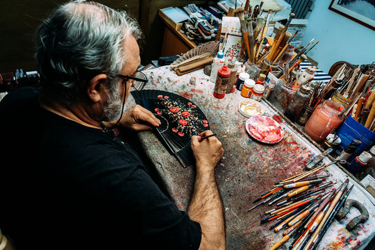 Painter using paintbrush to draw on hand fan while sitting at messy table and working in creative workshop