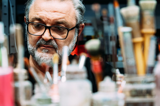 focused mature artist in eyeglasses working behind collection of paintbrushes of different sizes and bottles with paint in creative studio