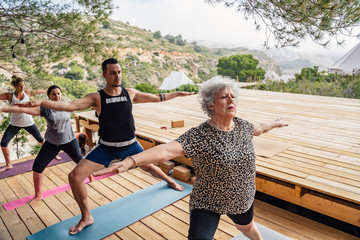 Group of motivated divers people in different ages stretching legs and ankles while performing Virabhadrasana B pose during outdoor yoga class