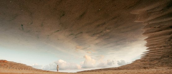 Full body anonymous person in beige casual clothes standing on sandy dunes in dry desert valley reflecting in cloudy sky mirage in desert