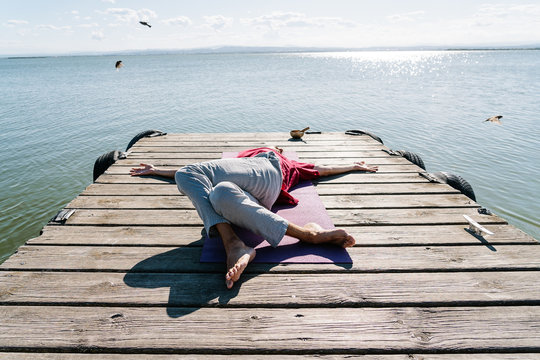 High angle of unrecognizable barefooted person lying on wooden pier against sea in Supta Parivrtta Garudasana pose during yoga session on sunny day