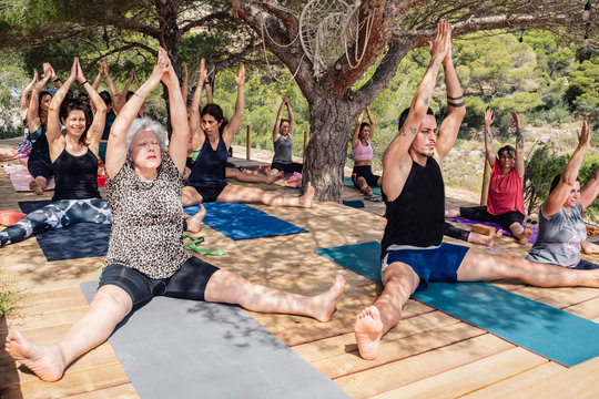 Group of diverse people of different ages doing Seated Wide Legged exercise with raised arms while practicing yoga in summer park