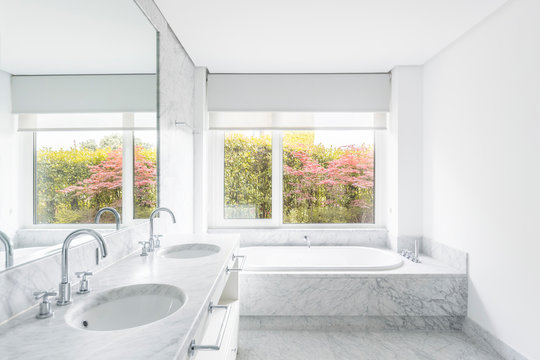 Creative minimalist design of bathroom with similar vanity units under big mirror and bath tube near window overlooking shrubs with blooming flowers