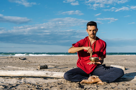 High angle of peaceful young barefooted male in casual clothes sitting on sandy beach in Half Lotus asana near waving ocean and playing Tibetan singing bowl against blue sky