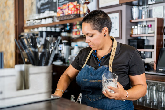 Positive focused female barkeeper in apron standing at counter making alcohol drinks and smiling while working in cafe