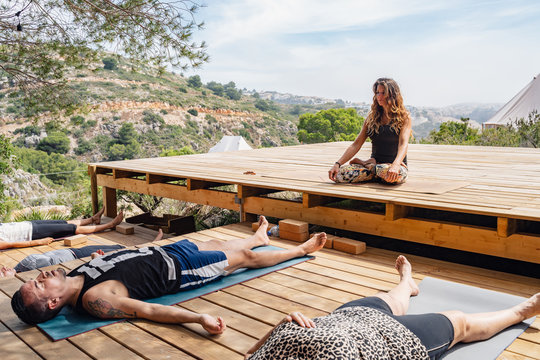 Female yoga trainer sitting in Lotus pose instructing students relaxing in Corpse asana after finishing outdoor workout on sunny day