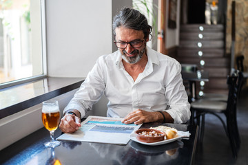 Positive ethnic bearded aged man sitting at table with of glass of foamy beer and reading newspaper near window