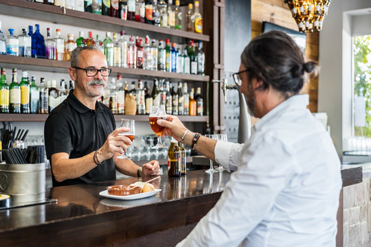 Aged man clinking glasses of bear with bearded barkeeper in black t shirt while having lunch in pub