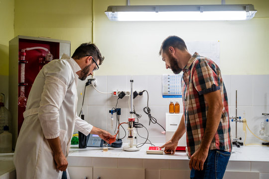 Side view of bearded chemist in eyeglasses standing near coworker in casual clothes in front of wine analysis equipment and flasks in lab
