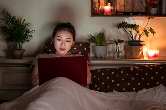 Young ethnic Asian female sitting on bed using netbook in the evening
