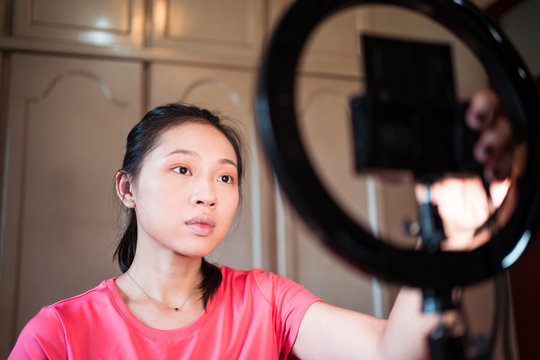 Focused young Asian woman stetting camera on tripod with ring light while preparing for creating new beauty blog at home