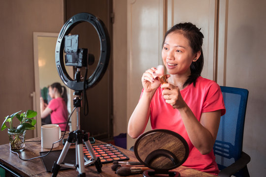 Cheerful young Asian female in casual outfit demonstrating bunch of various types of brushes for makeup while recording beauty video vlog at home