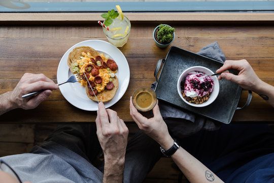 From above of tattooed woman eating muesli with natural yogurt decorated with blueberry jam and male partner eating delicious toasted bread with fried eggs and sausage slices with lemonade