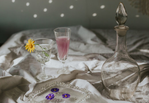 Above view of cocktail decorated with purple flower petals served on white tablecloth with vintage styled glasses and bottle on sunny day