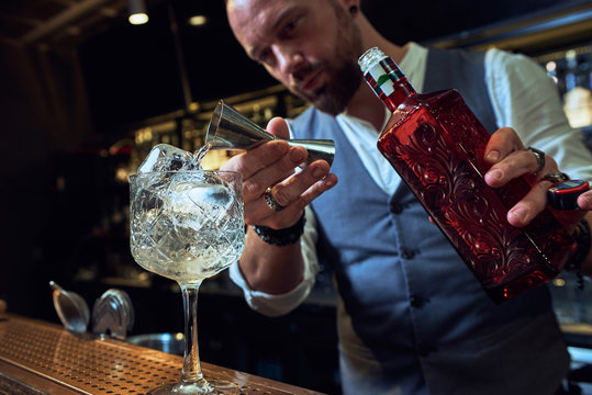 Professional young barman pouring alcohol cocktail from jigger into a glass on counter making beverage working in bar