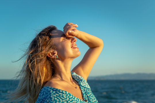 Side view of happy young female traveler in stylish outfit smiling with closed eyes while enjoying summer day on seashore against cloudless blue sky