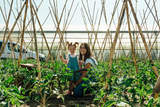 Cheerful woman cuddling adorable girl in denim sundress while squatting near small lush green tomato trees and wooden sticks in rural zone near van