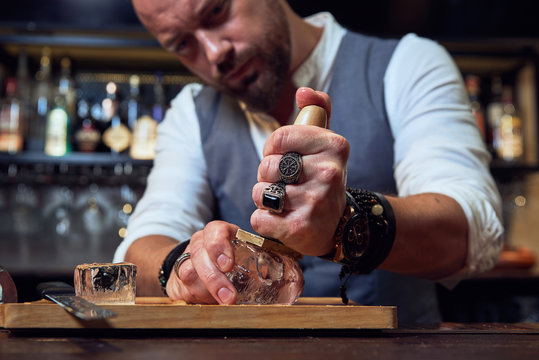 Professional bartender scratching ice cube with wooden tool on wooden board while preparing a cocktail in the bar