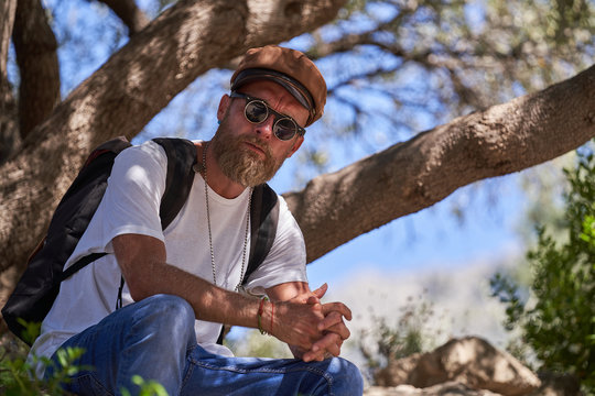 Low angle side view of contemplative adult hipster male traveler in cap and sunglasses with backpack relaxing under tree branches looking at camera during hiking in nature in sunny summer day