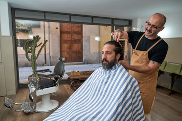 Barber combing hair while cutting hair to serious male customer in striped cape in modern barbershop with glass wall