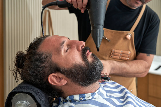 Unrecognizable crop barber drying beard with hairdryer and comb of man in chair with eyes closed