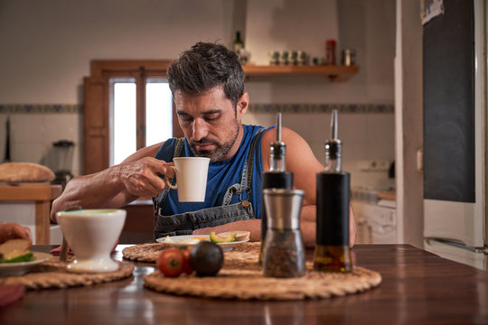 Calm male sitting at table in kitchen drinking fresh hot beverage in a mug and enjoying tasty breakfast