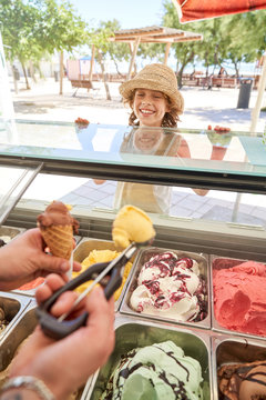 Crop ice cream shop assistant putting chocolate and vanilla ball in waffle cone for enthusiastic boy in straw hat