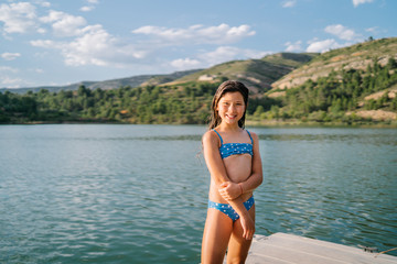 Happy little girl in bikini standing looking at camera in wooden pier near pond during summer holiday
