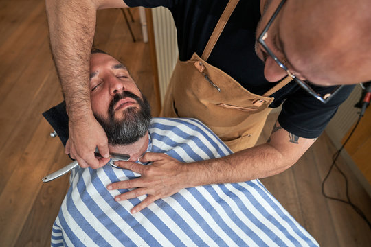 Barber doing beard hairstyling with sharp metal razor in barbershop