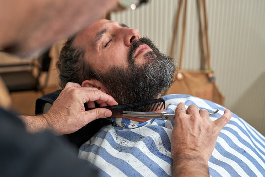 Hairdresser cutting beard of mature male client lying on comfortable chair in minimalistic studio