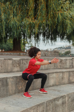 Side view of fit ethnic sportswoman in active wear and wearable bracelet doing squats using elastic tape while reaching arms on stairs under tree and listening to music from headphones
