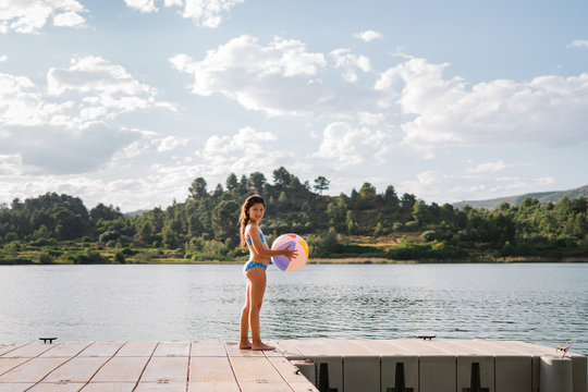 Side view of girl in bikini playing with beach ball while standing in wooden pier near pond during summer holiday