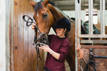 Content female equestrian in helmet and with bridle preparing chestnut horse for dressage while standing in barn