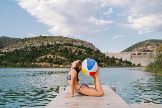 Side view of flexible girl in swimwear practicing yoga in Raja Bhujangasana with beach ball between head and feet