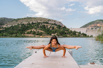 Smiling child in bikini doing yoga in Tittibhasana on quay near lake and looking at camera