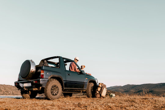Dreamy male traveler in hat and sunglasses sitting on auto near purebred dog with tongue out while admiring river and mountains on sunny day
