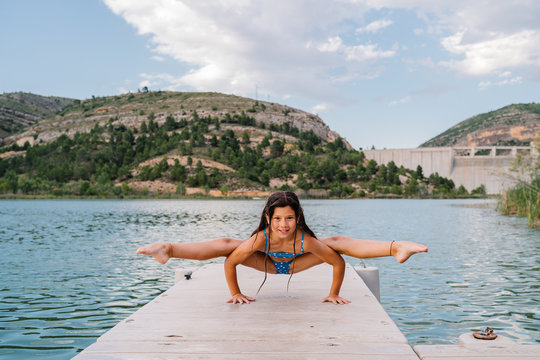 Smiling child in bikini doing yoga in Tittibhasana on quay near lake and looking at camera - Powered by Adobe